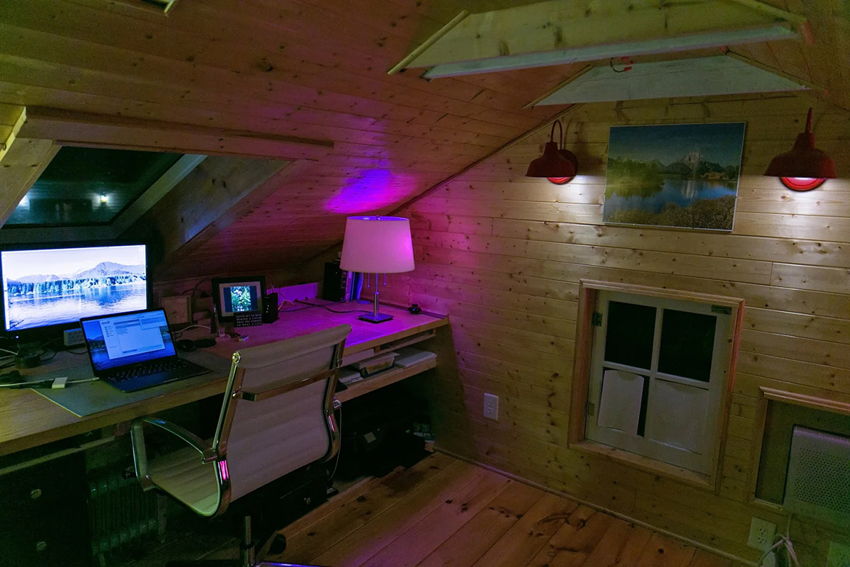 A wood finished attic room with a desk on the left with a chair and computer and purple lighted lamp