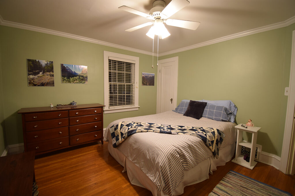 A bedroom with two dressers on the left, a window, door way, bed and side table to the left. There is a ceiling fan fixture lighting up the room