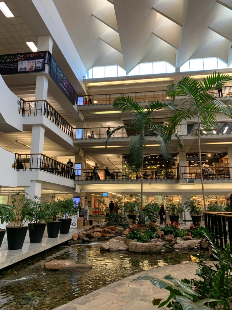 An indoor court at Dallas Market Center filled with plants and two palm trees and a sizeable pond. People are milling about and standing on balconies on three levels of the mall