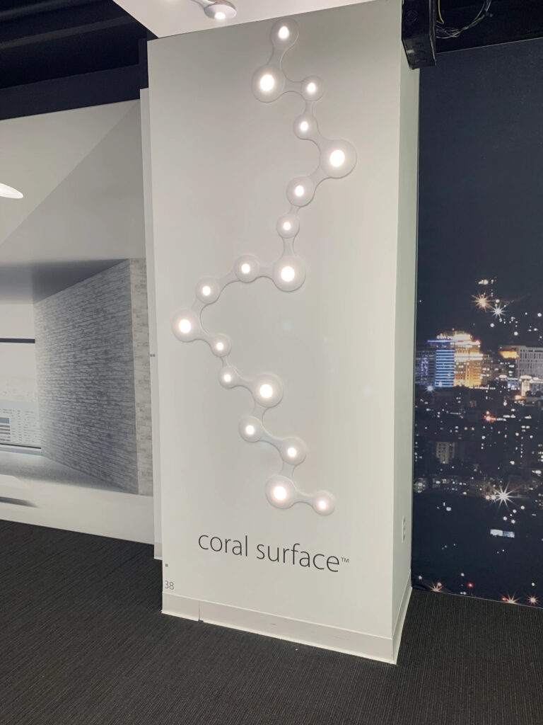 A small display wall that is situated next to a window with a view down onto the lights of a city at night. The display is modular, connectable round surface lights labelled "coral surface"