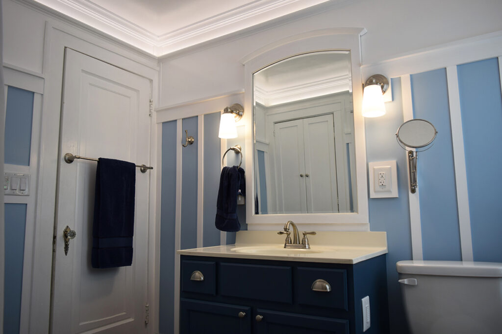 A bathroom sink with a mirror above it and two light on either side of the mirror and a small mirror hanging to the right. A towel rack and a door are on the left.