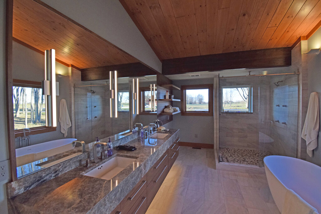 A very expensive looking modern bathroom with a wood ceiling and marble counters and floors. There is a glass shower on the right. The counter on the left has two sinks and a full wall mirror with lights attached to it. There is a window in the back. 
