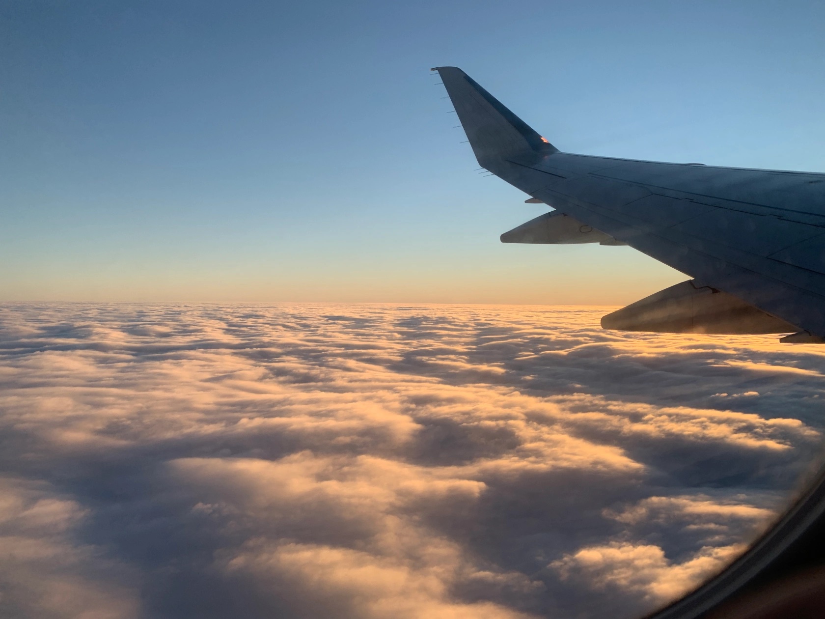 A photograph taken outside of an airborne airplane during golden hour right before sunset. The photo features the wing of the plane on the right, which is also where the sunlight is coming from. There is a sea of clouds along the bottom, with the horizon at about halfway up the photo. The clouds glow slightly golden. The plane wing is mostly in shadow.
