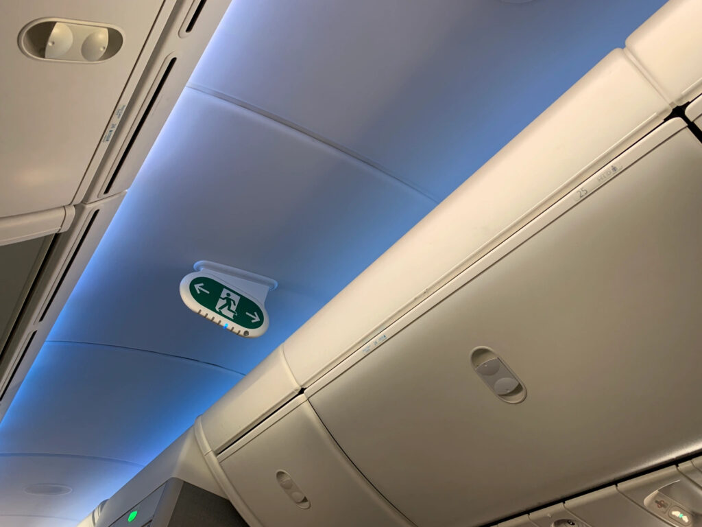 The ceiling of an airplane and part of the overhead storage. There is a small exit sign with an icon of a person running and an arrow on the left pointing left and an arrow on the right pointing right. 