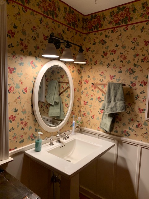 A bathroom pedestal sink and oval mirror with three lights pointed down on the sink. There is yellow and red floral wall paper, white wainscotting, and a towel on a rack to the right.