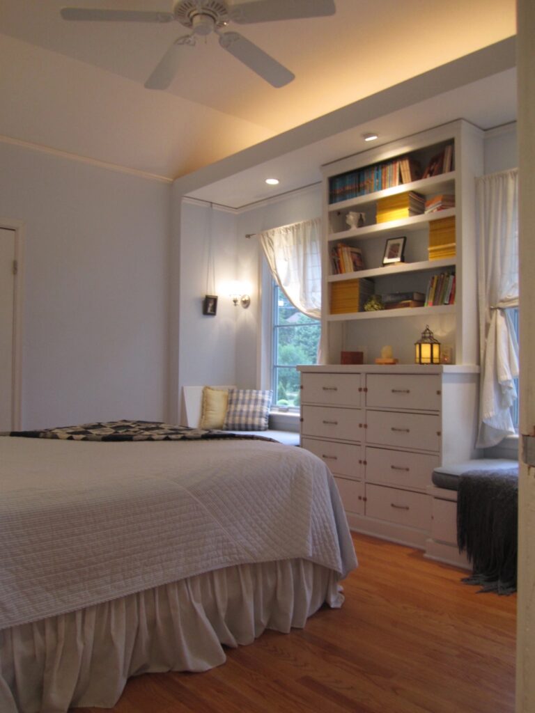 A photograph of a bedroom with white walls, white bedspread, and white shelving unit across the room from the bed. There is cove lighting above the white shelf and circular lights that illuminate parts of the shelf and windows on either side of the shelf.