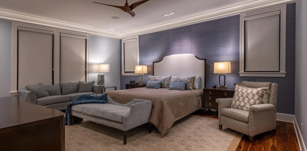 A photograph of a furnished bedroom with a bed in the middle, a couch on one side and an easy chair on the other. There are three lamps, two on either side of the bed and one on the couch. Where the ceiling meets the wall, there is cove lighting illuminating the space.