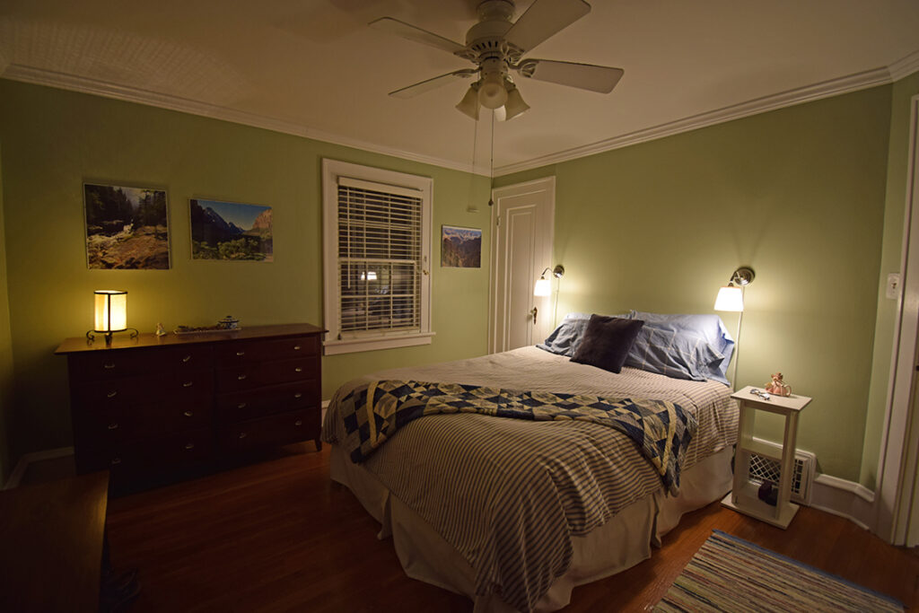A bedroom with a ceiling fan, several large pieces of art on the wall, and a low wide chest of drawers with a lamp on it. There are two lamps on either side of the bed.