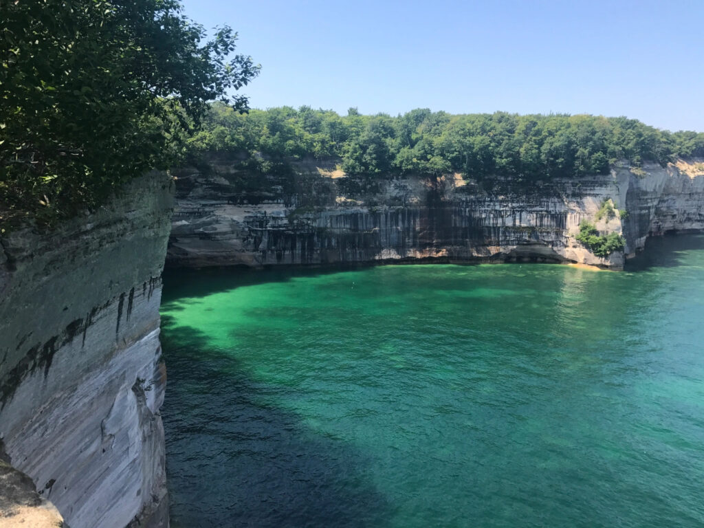 Sheer rock cliffs with trees atop them on the left and very blue-green water below opening to the right, on a sunny day.