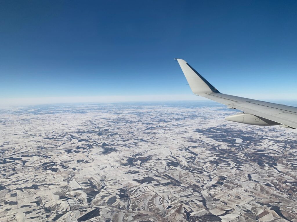 Photograph of a view from an airplane window. The plane's wing is in the right side of the photograph. The earth below extends to the horizon, about halfway up the photo. The land below is covered in snow. The sky is bright blue and cloudless.