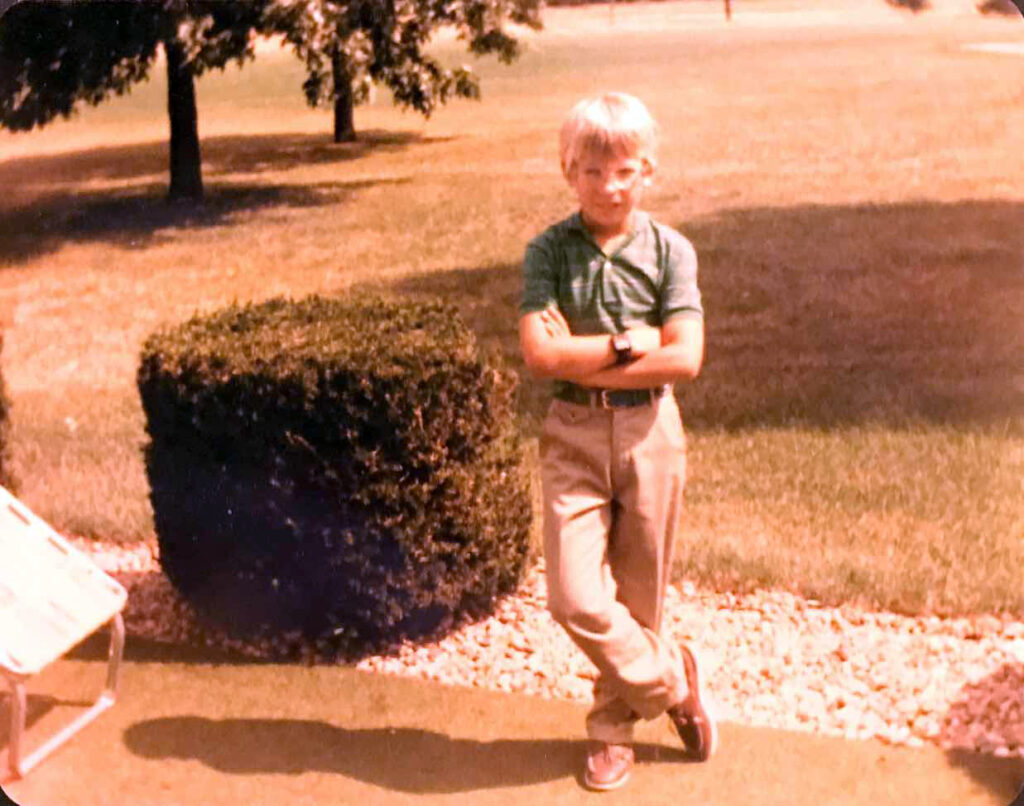 A kid in the 70s standing in front of a bush with his arms crossed