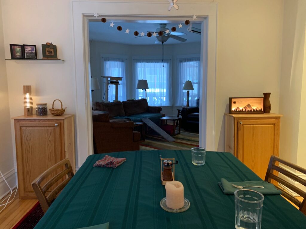 A photo of a dining room table with a green tablecloth. Beyond the table is a living room cast mostly in shadow with natural light from three curtained windows. The dining room is lit by a single light coming from the right off-camera, plus two lamps on cabinets standing on either side of the doorway. 