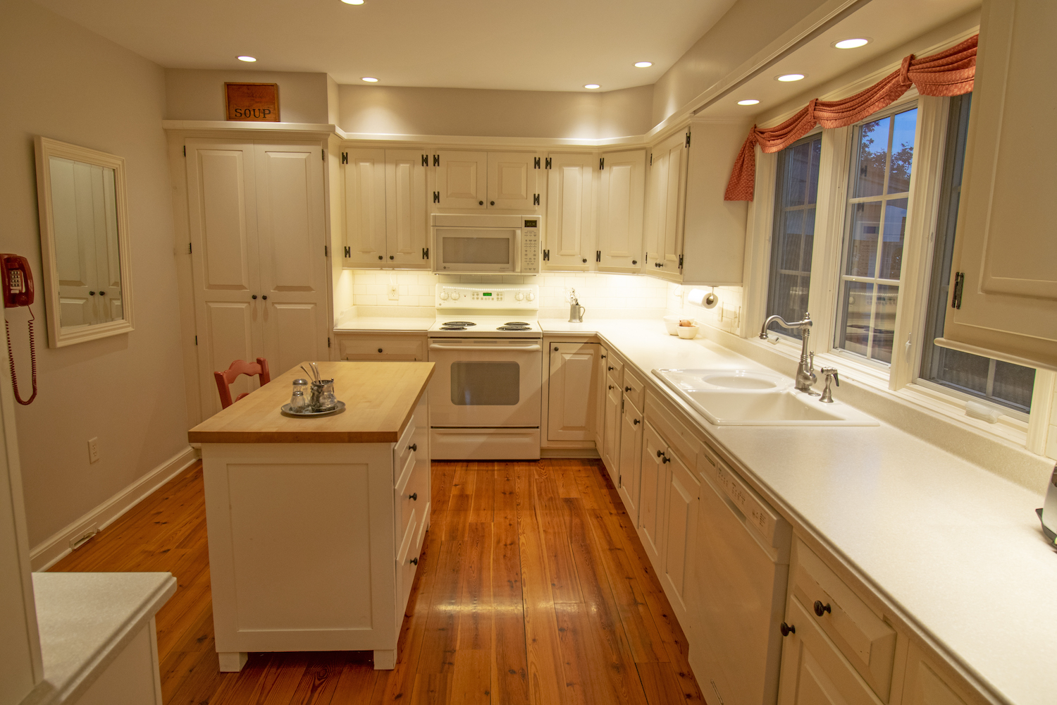 A photograph of a kitchen with white cabinets, walls, and countertops, and nice hardwood floor. There is lighting underneath the cabinets, circle lights in the ceiling, and everything is very well lit.
