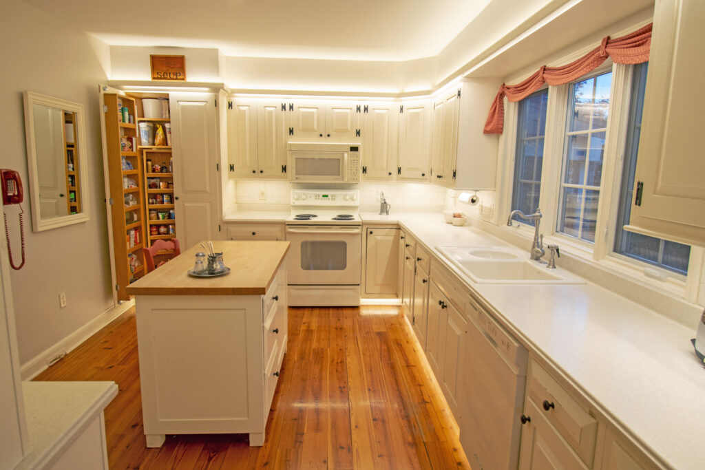 A photograph of a kitchen with white cabinets, walls, and countertops, and nice hardwood floor. There is lighting underneath the cabinets, along a grove over the cabinets, and underneath the counters by the basebords. Everything is very well lit.