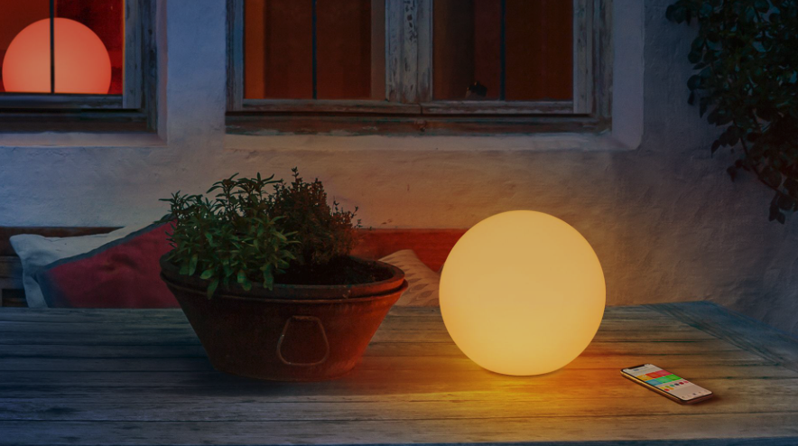 An outdoor wooden table in front of a wall with windows, with a potted plant and globular light atop it, at night