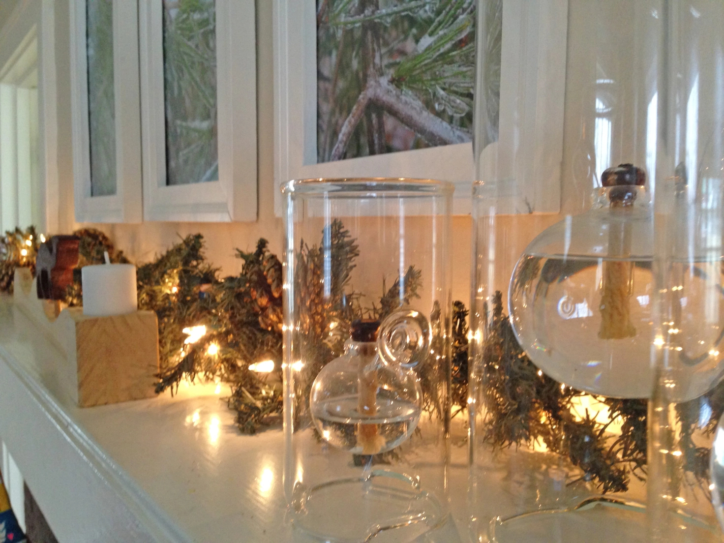 A mantle with three photos of icy pines above it. There is a long garland of fake pine and holiday lights along the top behind several glass lanterns and a wooden candle holder with a camel