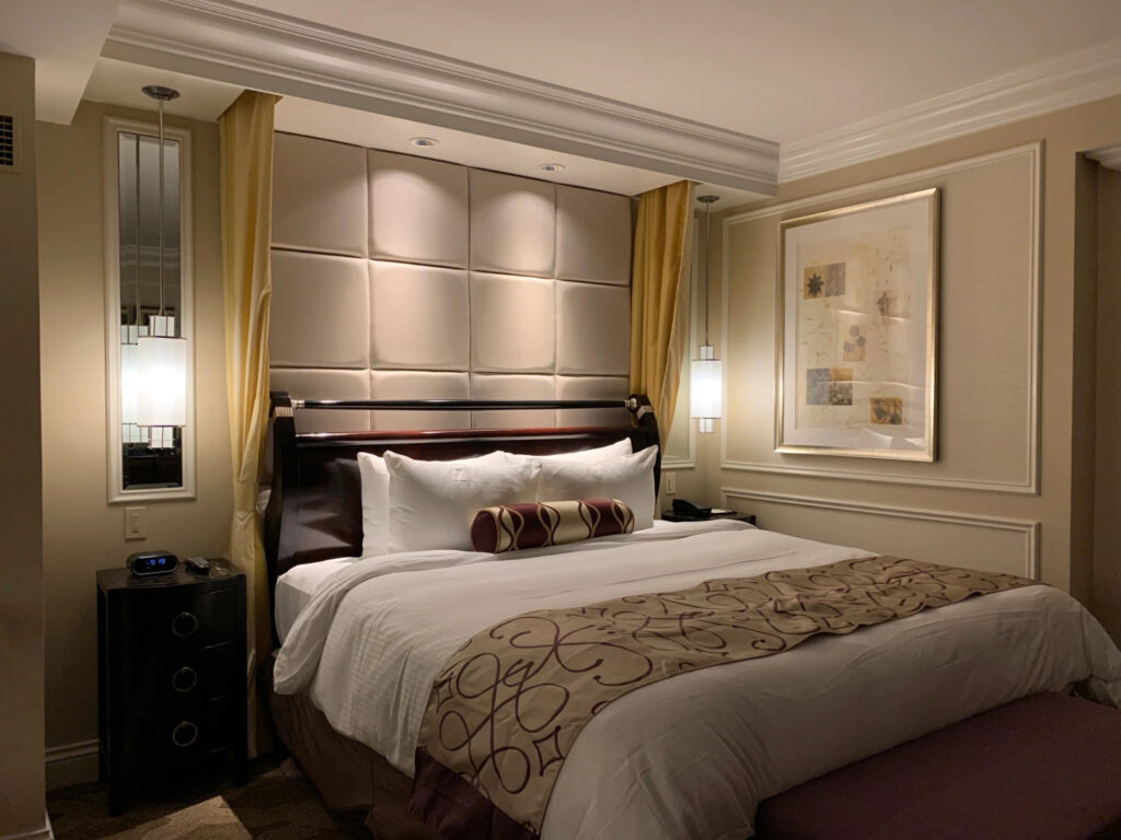 A fancy hotel bedroom with two wall sconces in front of mirrors on either side of the bed. there is a piece of art hanging to the right of the bed, and two small bedside tables on either side of the bed.