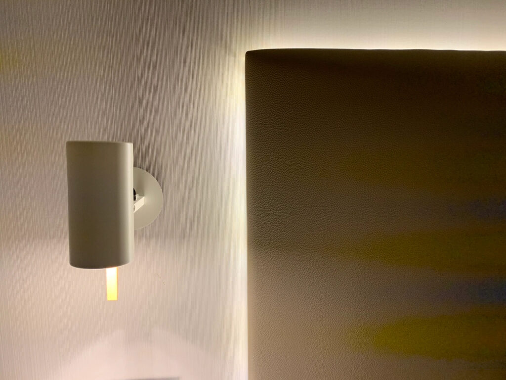A white cylindrical bedside downlight next to a simple rectangular headboard that is lit from behind by LEDs