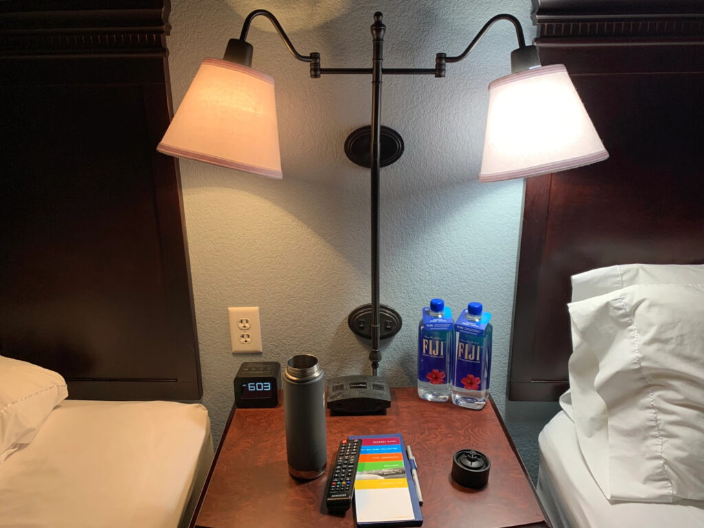 The bedside table in a hotel room with two lamps pointing at beds on either side of it.