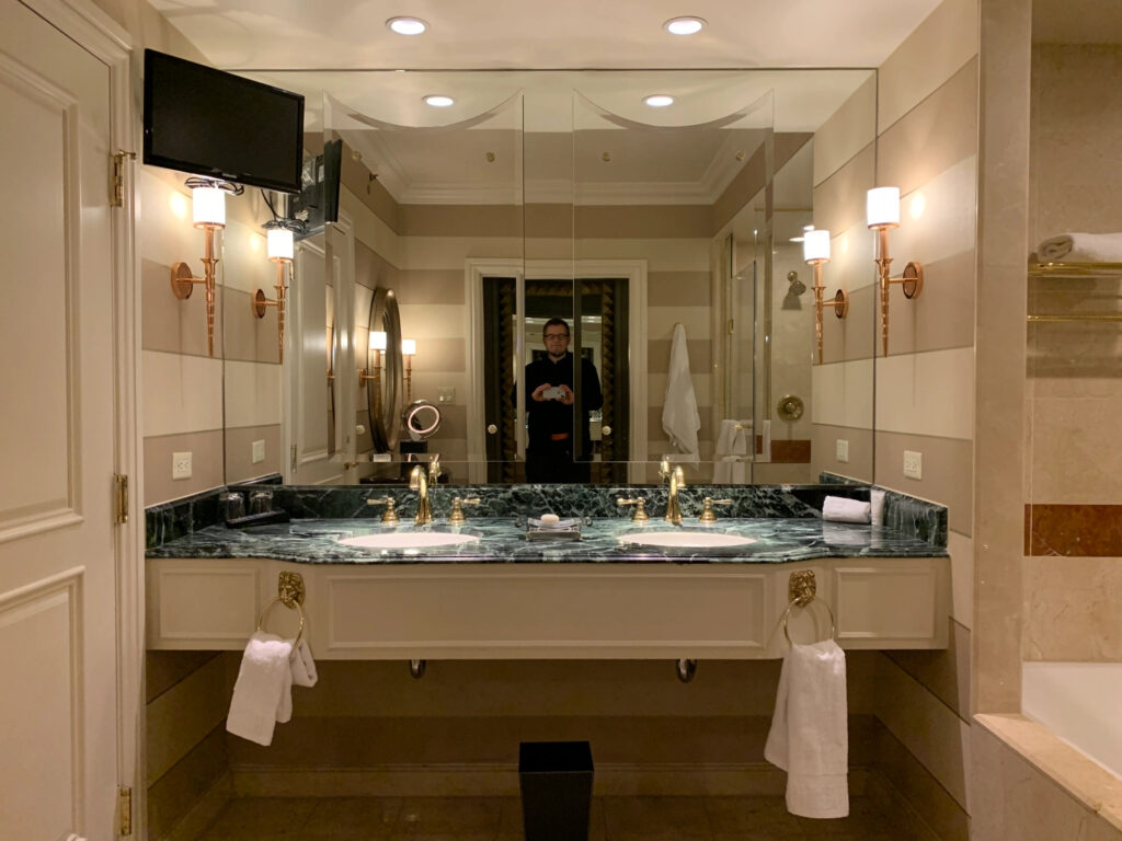 A fancy hotel bathroom with a wall-to-wall, sink to ceiling mirror, two sinks with a marble top, a tv screen in the upper left, and two wall sconces on either side. A bath is to the right and a door to the left. There is a man taking a photo in the mirror.