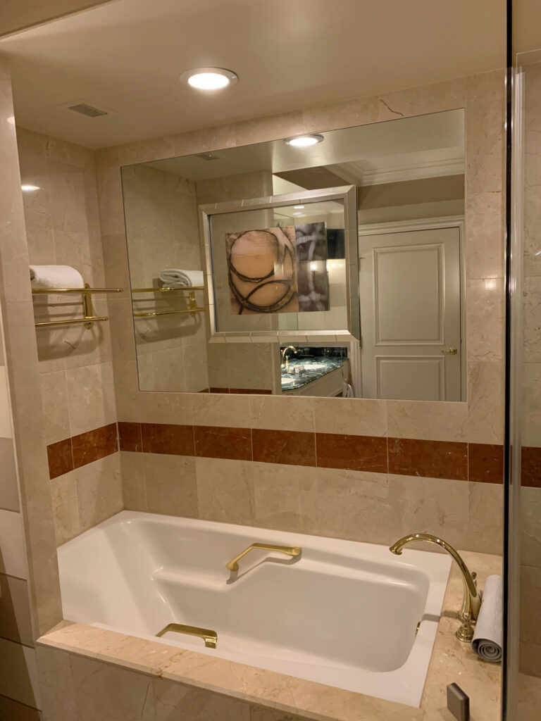 A fancy hotel bathtub with a towel rack at the foot of the tub and a large mirror with a piece of art in the middle of it to the side of the tub.
