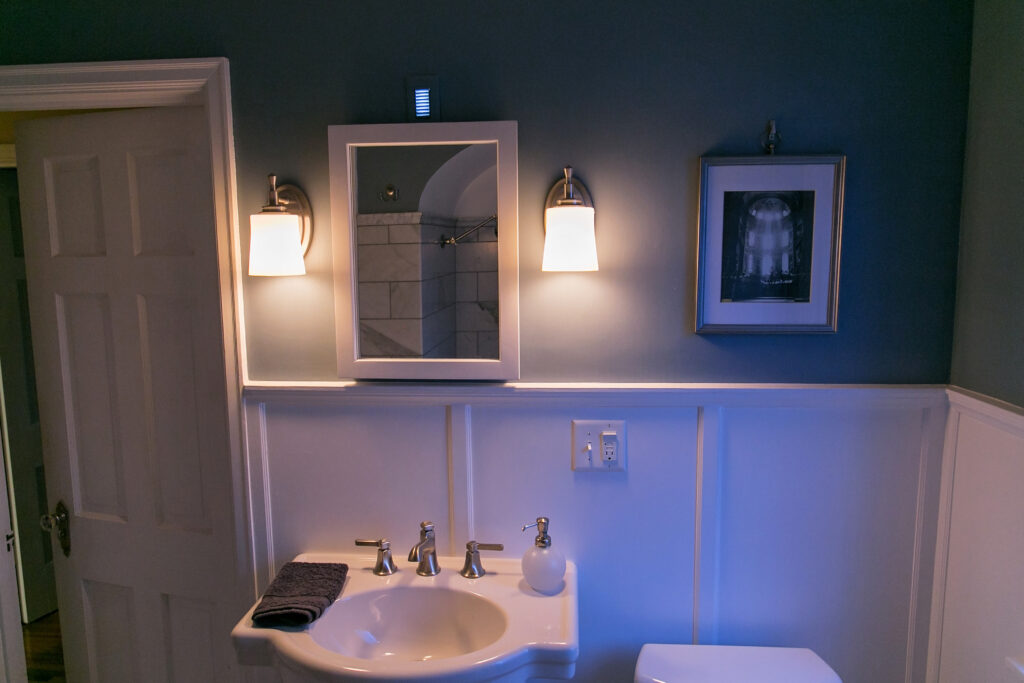 A photograph of a bathroom with white sink and mirror over the sink. There are two wall sconces on either side of the mirror, both lights are turned on.