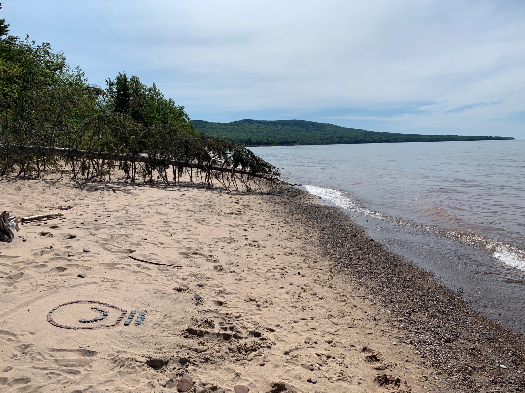 A photograph of a sandy beach along a lake shore. Someone drew the Light Can Help You logo in the sand.