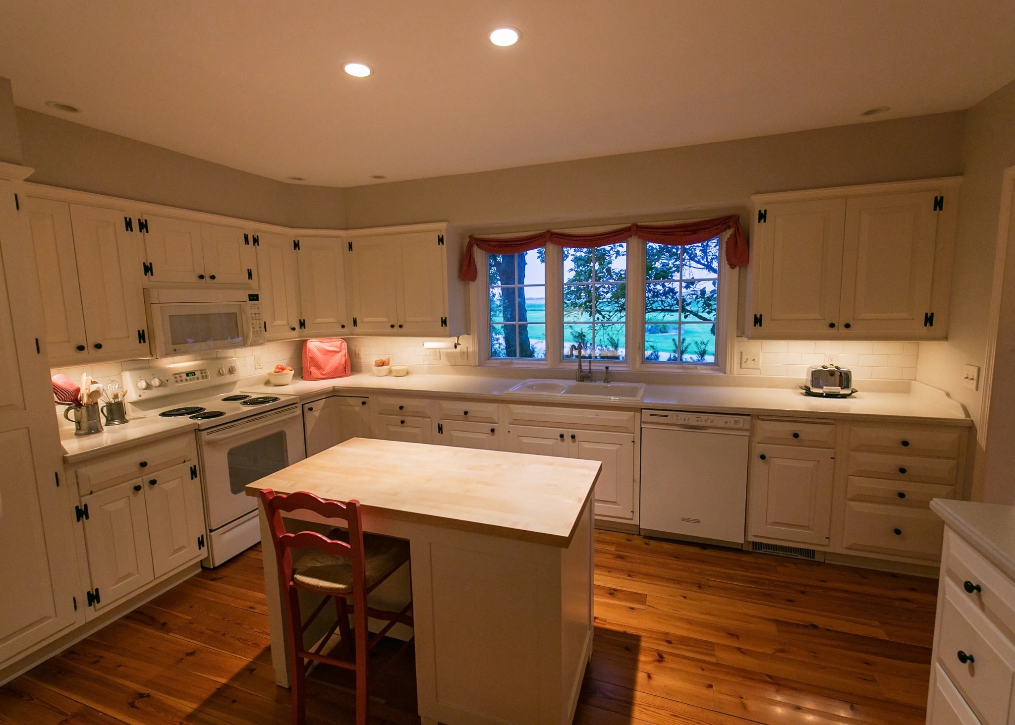A photograph of a well-lit kitchen featuring lights underneath the cabinets and ceiling lights directly over the island. The cabinets are white with dark handles and the floor is hardwood.