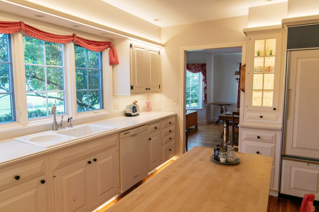 A white kitchen with a large window above the sink with a view of trees, a butcher block island in the center, and a doorway to a dining room to the right of the sink