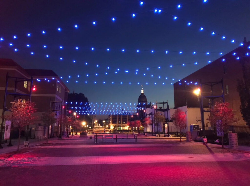 A view of Library Mall at the end of State Street in Madison WI facing the state capitol. There are blue fairy lights strung up above the whole square with pink spot lights lighting up the cobblestone