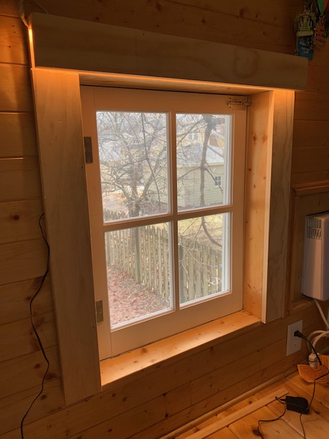 A window with four panes in a wooden wall lit from above with tape LED lights, view of the outdoors on a sunny day