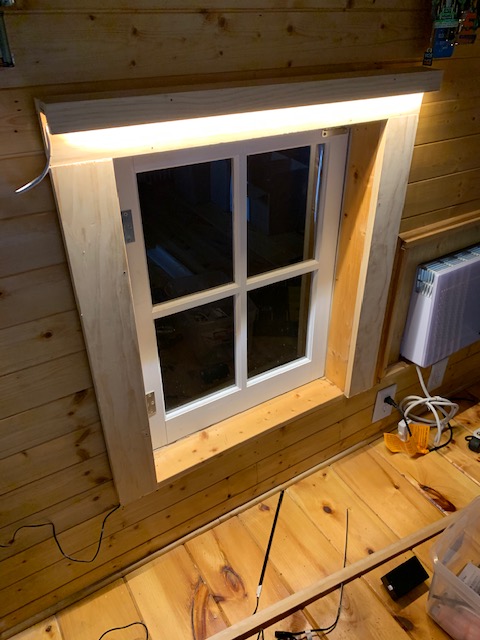 A window with four panes in a wooden wall, lit from above with tape LED lights, it's dark outside
