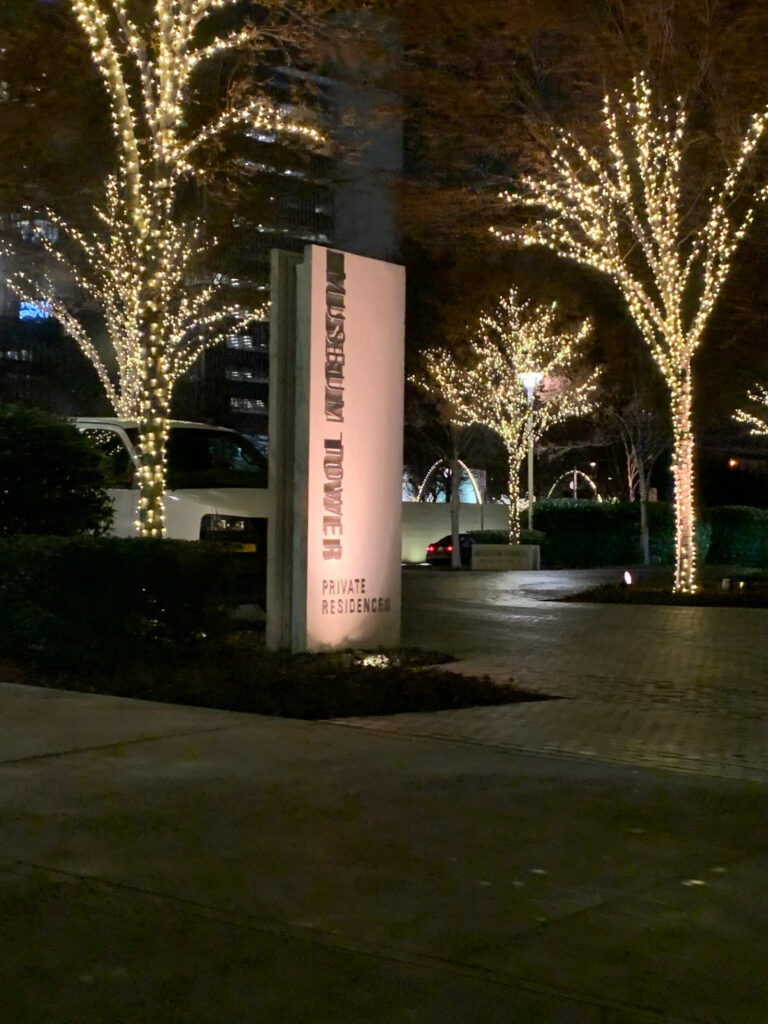 The entrance to a private apartments courtyard at night. The sign is lit up from underneath. The trees are covered completely in twinkle lights. 