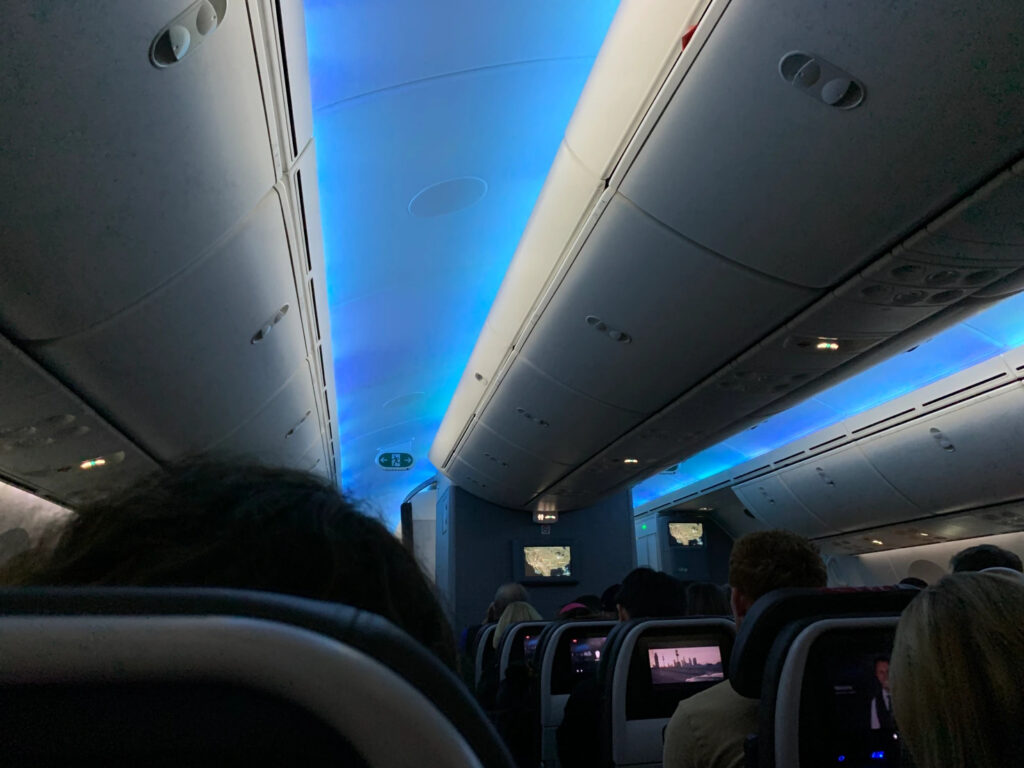 Airplane passenger area at night. The ceiling is lit up blue, everyone has a screen on the back of the seat ahead of them.