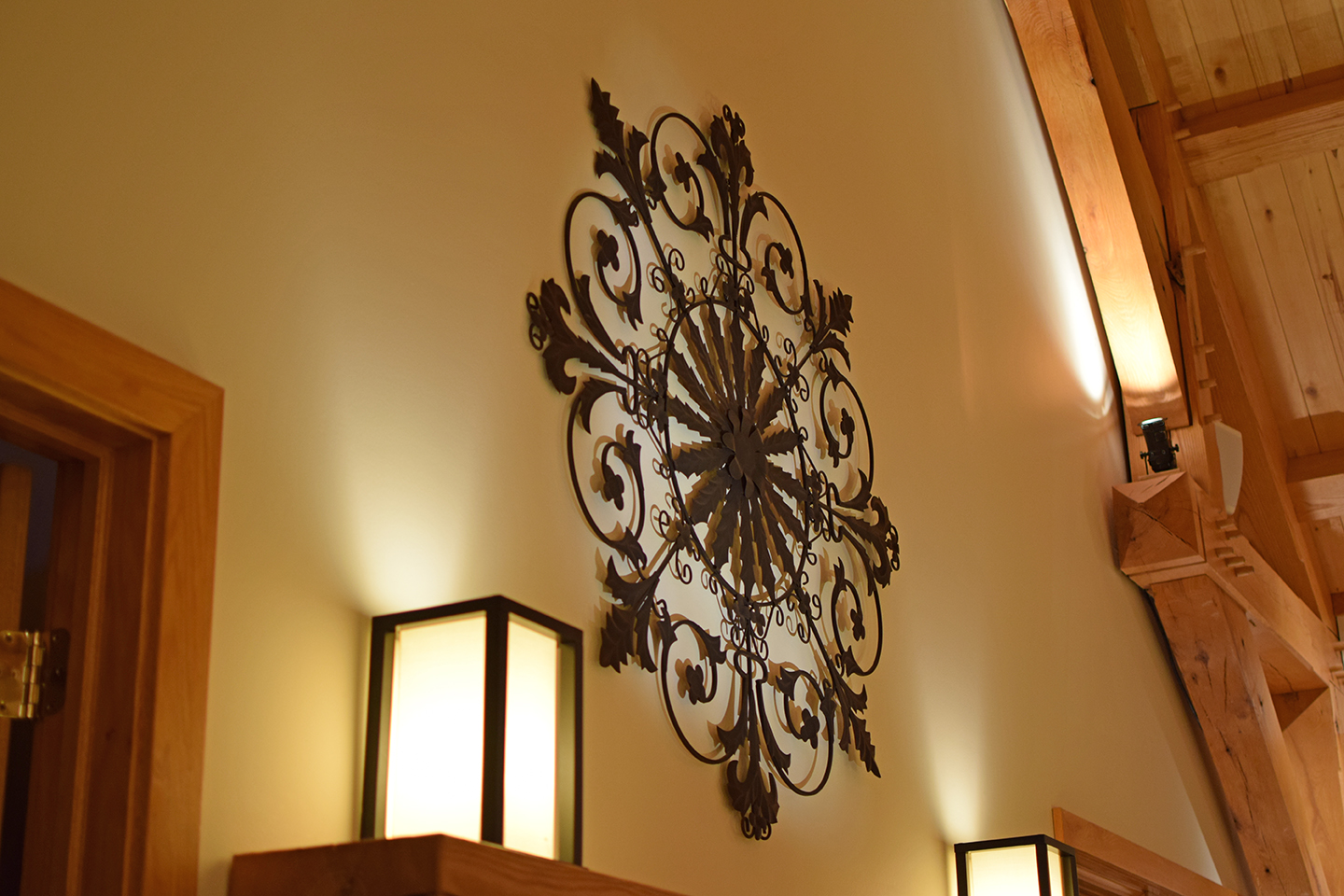 A wall in a home with a large, ornate, flat metal sculpture on the wall with two lamps below on either side.