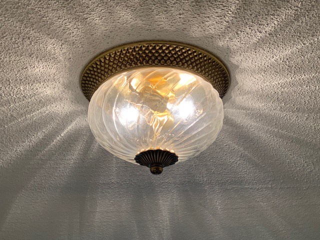 An overhead light that has a gold base and tip, and a textured clear glass globe with the light turned on