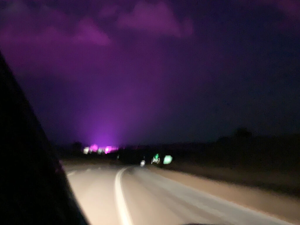 The road at night from a moving car. There are purple lights in the distance.