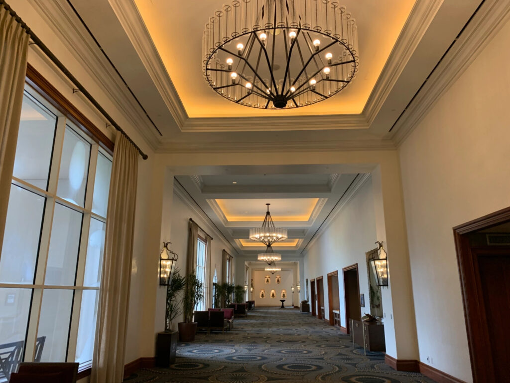 A wide hallway in a resort hotel with windows and potted palms on the left and doorways on the right, with four chandeliers at intervals