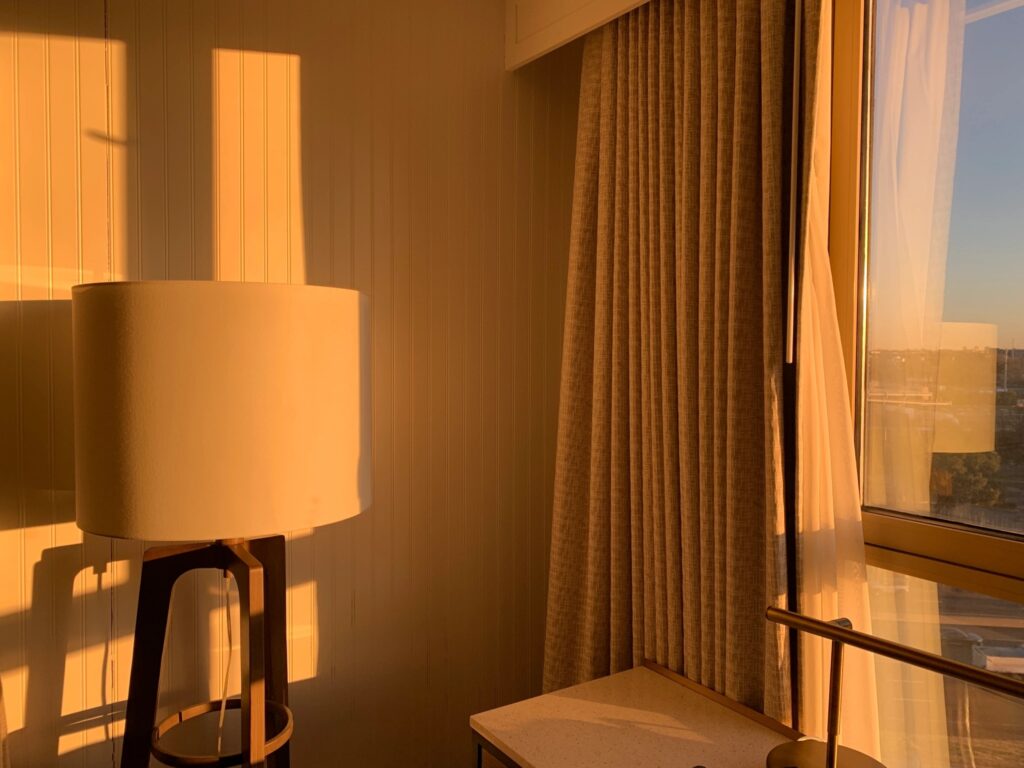 Photograph of a corner in a hotel room. The right side is a window with the curtains open. On the right is a floor lamp with lampshade. The setting sun colors everything with a golden gleam.