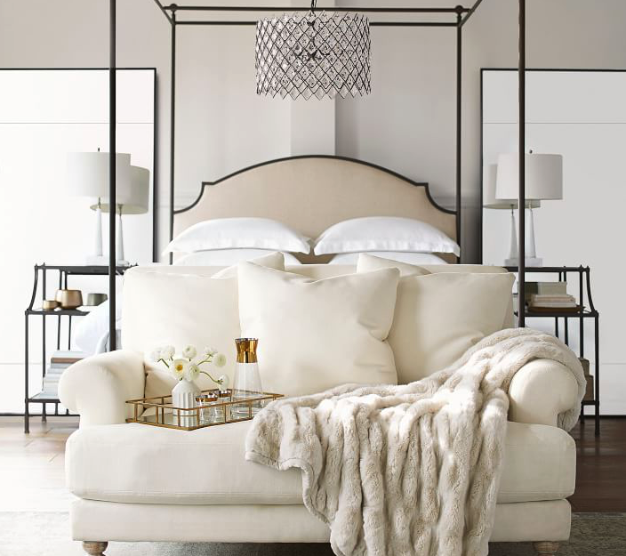 Photograph of a white loveseat at the foot of a bed. The walls, loveseat, and bedspread are white with a dark bedframe and four posters.