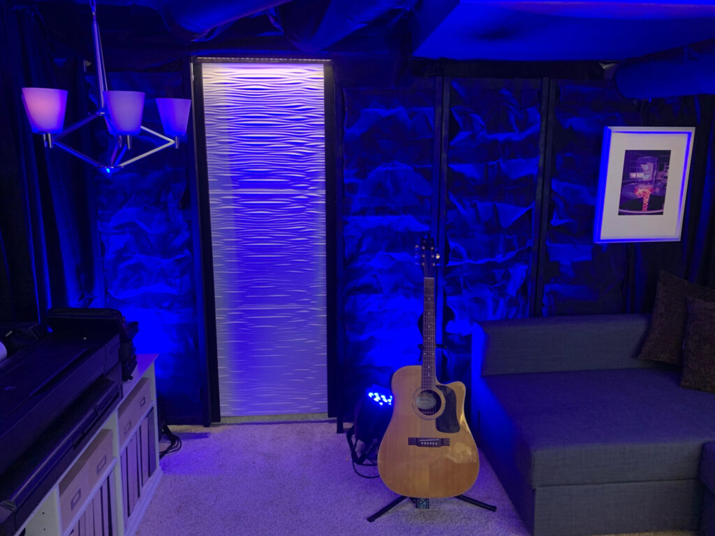 A room lit by blue light. There's a table with storage under it to the left, an acoustic guitar, painting and couch to the right. There is a doorway in the middle.