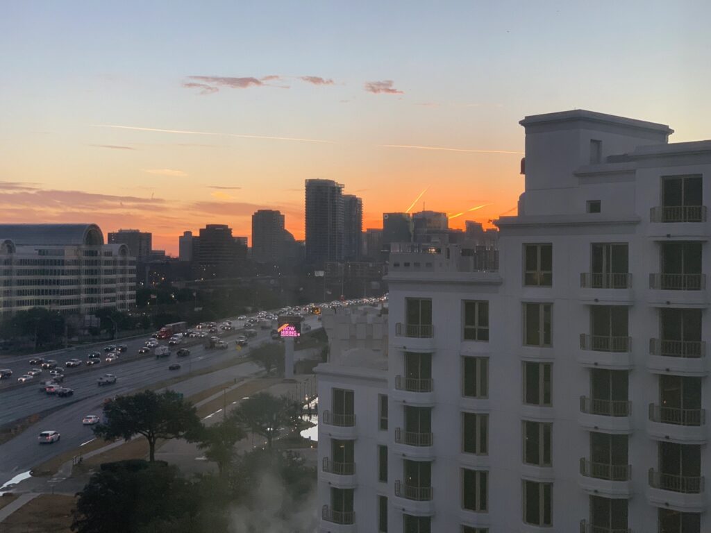 Photograph taken of the view of a hotel window. To the right is a white building. To the left is a busy highway. The horizon is a city's downtown, with a glowing orange sunrise right behind that.