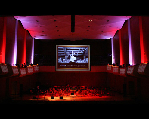 A theater view of the stage with only the stage and pillars lit with red and purple light, the stage is covered in chairs. Above that is a black and white video playing on a large screen.