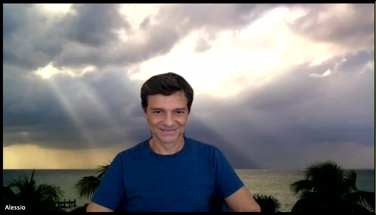 A man with a background green-screened behind him of an outside view of the ocean on a dark cloudy day with some trees peeking up on the bottom. Light is filtering through the clouds in rays.