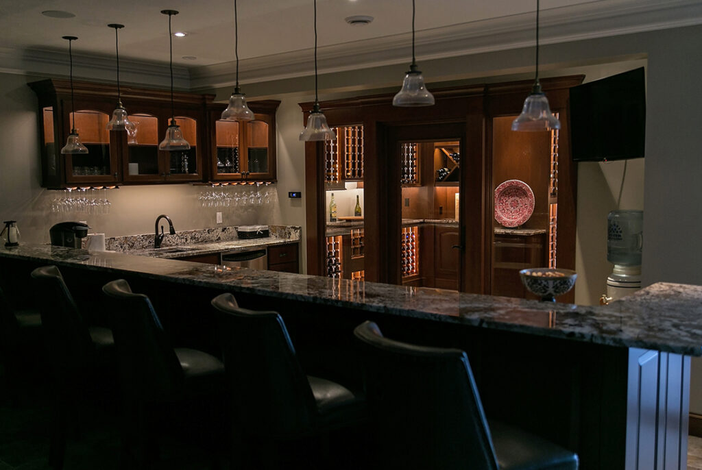 A photograph of a kitchen with a long countertop for diners. The only lights on are the ceiling lights and the lights underneath the cabinets. It looks very dark.
