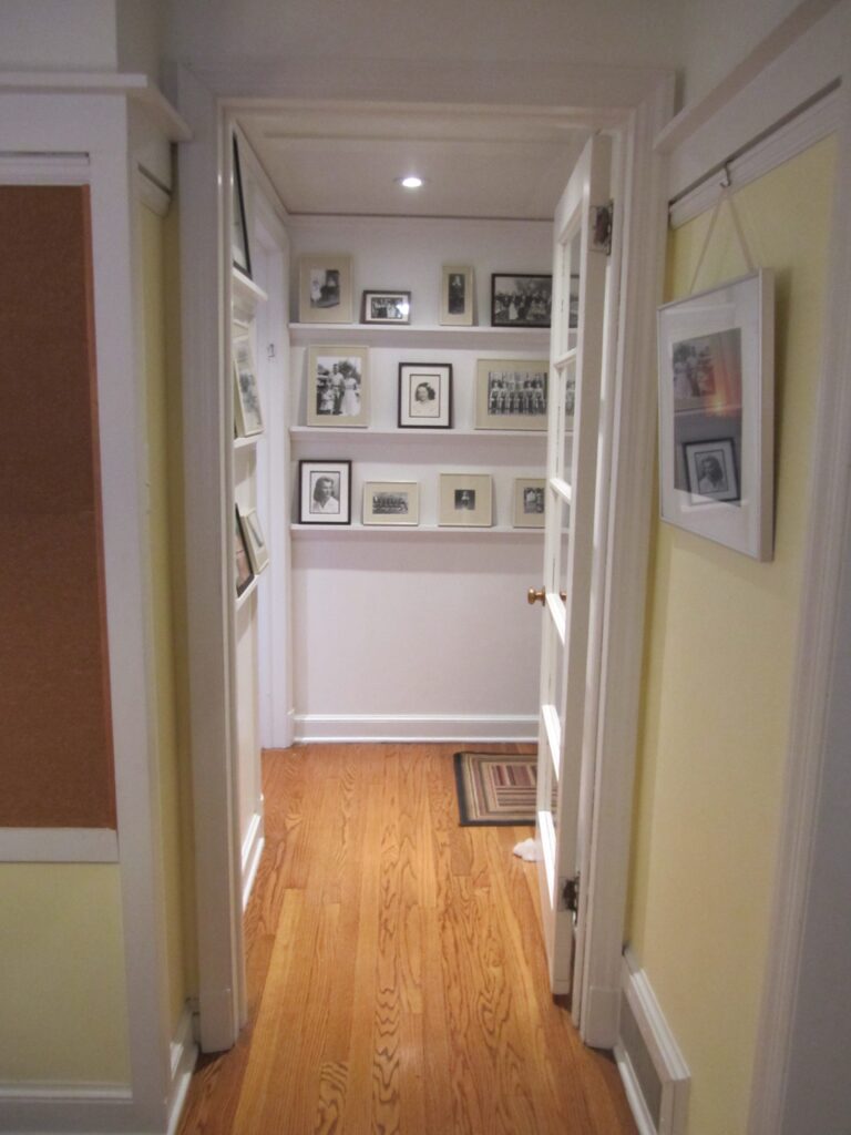 A photograph of a hallway with white walls and thin, white shelves holding up a series of picture frames. The floor is hardwood. 