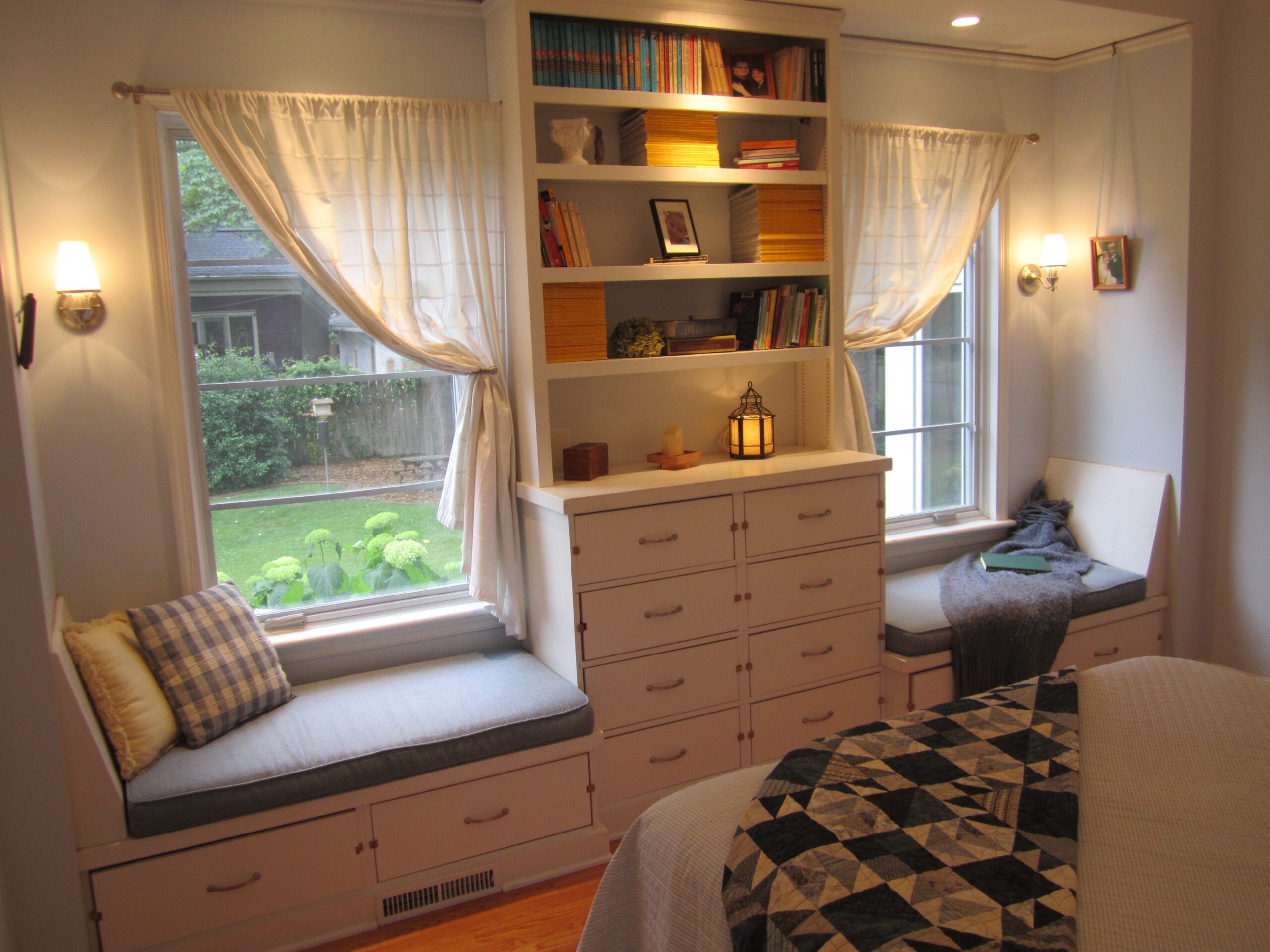 A photograph of a white bedroom with two windows and a bookshelf between the windows. The nooks by the windows are lit by wall sconces