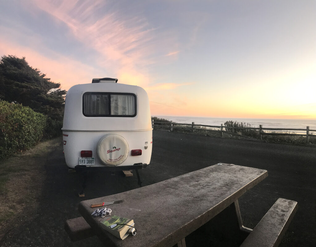A photograph of a camper by a picnic table. Overhead is a pink-and-blue sky, signifying a sunset.